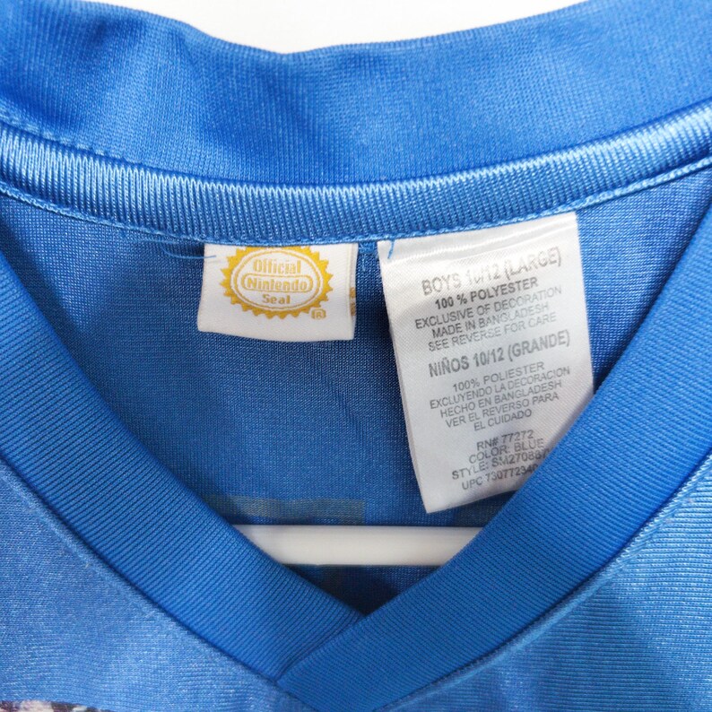 Puede incluir: Una camiseta azul con cuello en V que presenta una etiqueta Official Nintendo Seal y una etiqueta de cuidado blanca. La etiqueta especifica 100% poli&eacute;ster, talla 10/12 Large, e indica que la prenda fue fabricada en Bangladesh. Textura sint&eacute;tica lisa.