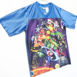 Puede incluir: Una camiseta azul con cuello en V que presenta un estampado gr&aacute;fico vibrante de Super Mario Galaxy. El dise&ntilde;o muestra varios personajes, incluidos Mario, una planta Pira&ntilde;a y Bowser sobre un fondo espacial morado oscuro con un planeta naranja brillante.