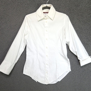 May include: A crisp white long-sleeve button-down shirt featuring a structured collar, a central vertical button placket, and tailored seams along the front. The sleeves are finished with wide cuffs, and the hem has a curved shape.