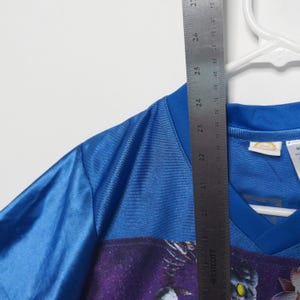 Puede incluir: Una camiseta deportiva azul con cuello en V y un estampado gr&aacute;fico con un fondo espacial morado y una criatura. Una regla met&aacute;lica Westcott est&aacute; colocada verticalmente sobre la prenda, mostrando una longitud de unos 68,5 cm.