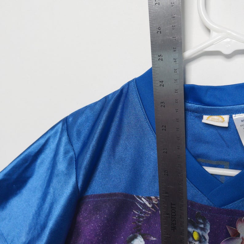Puede incluir: Una camiseta deportiva azul con cuello en V y un estampado gr&aacute;fico con un fondo espacial morado y una criatura. Una regla met&aacute;lica Westcott est&aacute; colocada verticalmente sobre la prenda, mostrando una longitud de unos 68,5 cm.