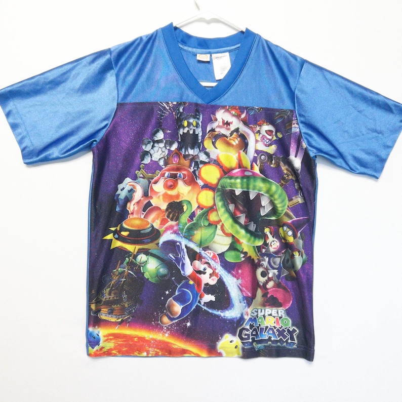 Puede incluir: Una camiseta de manga corta con cuello en V que presenta un estampado gr&aacute;fico vibrante de Super Mario Galaxy. El dise&ntilde;o muestra varios personajes, incluidos Mario, Bowser y una Planta Pira&ntilde;a sobre un fondo espacial morado con un planeta.
