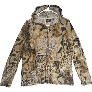 Puede incluir: Una sudadera con capucha y cremallera de manga larga con un estampado animal en tonos marrón, beige y negro. La prenda incluye capucha, cierre de cremallera frontal y un adorno central de pequeños remaches plateados. La etiqueta interior dice Opque.