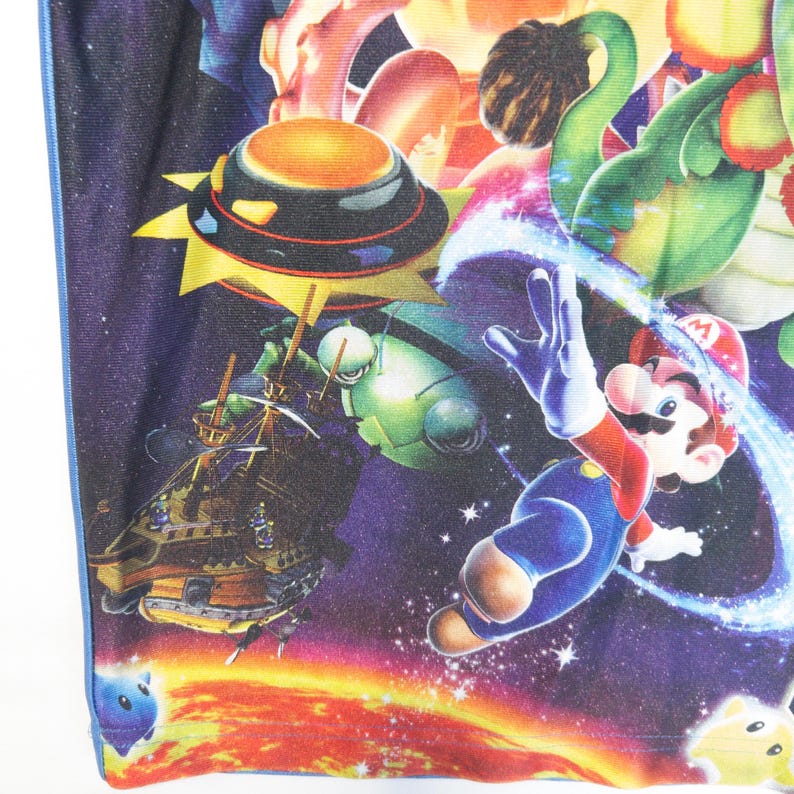 Puede incluir: Una prenda de tela con un estampado gr&aacute;fico vibrante de tem&aacute;tica espacial de Mario de Super Mario Galaxy. El dise&ntilde;o incluye a Mario flotando en una nebulosa p&uacute;rpura estrellada, un dirigible de madera, un personaje rob&oacute;tico verde y un sol.
