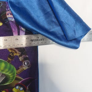 Puede incluir: Una camiseta con mangas azul brillante y un estampado gr&aacute;fico de tem&aacute;tica espacial en color morado que muestra un personaje. Una regla met&aacute;lica Westcott se coloca sobre la prenda, mostrando una medida de unos 46 cm en el pecho.