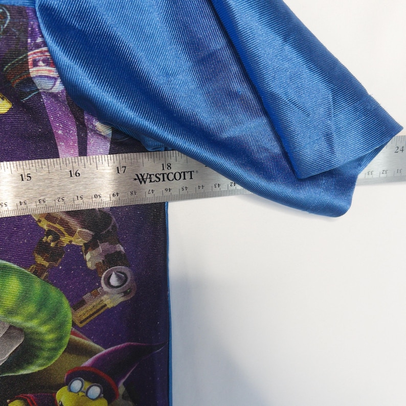 Puede incluir: Una camiseta con mangas azul brillante y un estampado gr&aacute;fico de tem&aacute;tica espacial en color morado que muestra un personaje. Una regla met&aacute;lica Westcott se coloca sobre la prenda, mostrando una medida de unos 46 cm en el pecho.