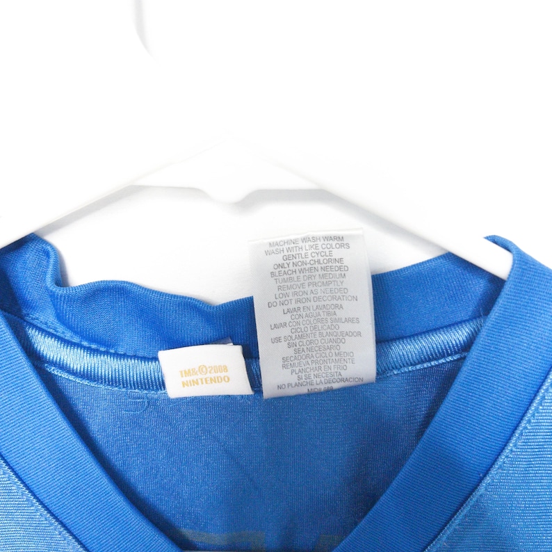 Puede incluir: Una camiseta azul brillante con cuello redondo acanalado y una etiqueta de cuidado blanca en el interior con instrucciones en ingl&eacute;s y espa&ntilde;ol. Una etiqueta blanca m&aacute;s peque&ntilde;a muestra el texto TMK 2008 NINTENDO. La prenda est&aacute; en una percha blanca.