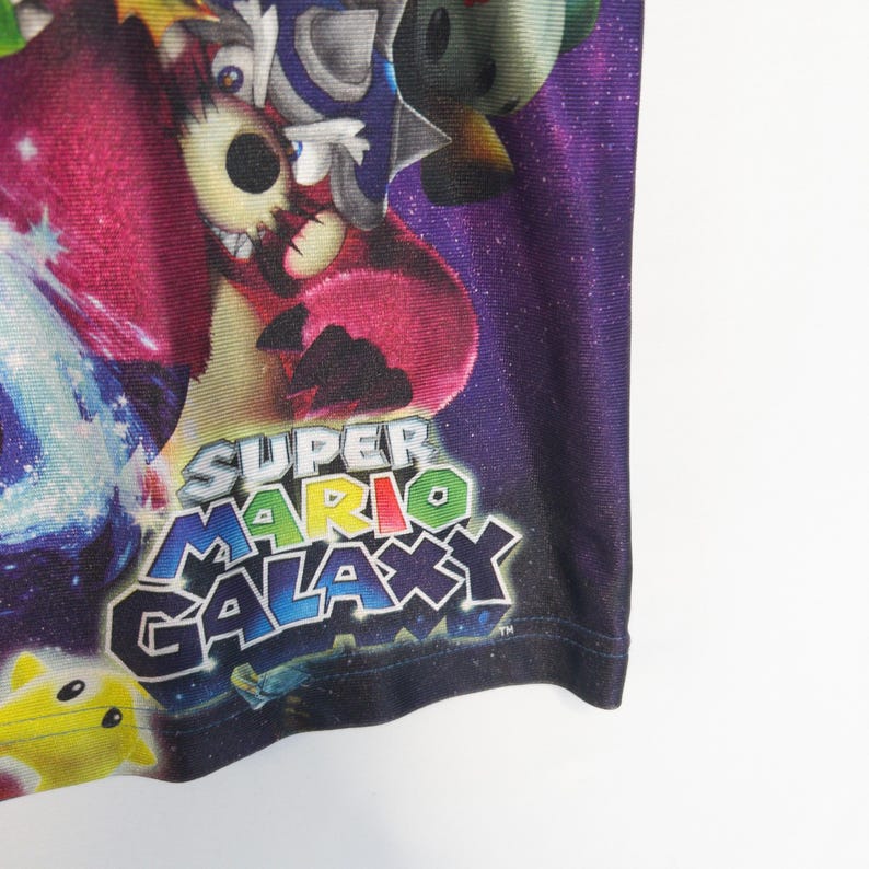 Puede incluir: Una prenda de tela morada y negra con un estampado gr&aacute;fico vibrante de personajes del videojuego Super Mario Galaxy. El dise&ntilde;o incluye un fondo con tem&aacute;tica espacial con estrellas y el logotipo de Super Mario Galaxy en letras.