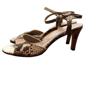 Op de afbeelding: Een paar sandalen met open teen en een slangenleerpatroon in bruin-, beige- en crèmetinten. De sandalen hebben een dun enkelbandje met een zilverkleurige metalen gesp en een hak van gemiddelde hoogte. De binnenzool toont de merknaam AGL.