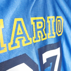 Puede incluir: Una camiseta deportiva de color azul brillante con el texto amarillo "ARIO" en fuente de bloque. Debajo del texto, un n&uacute;mero "7" grande de color blanco est&aacute; impreso en la tela. El material tiene una textura acanalada horizontal visible.