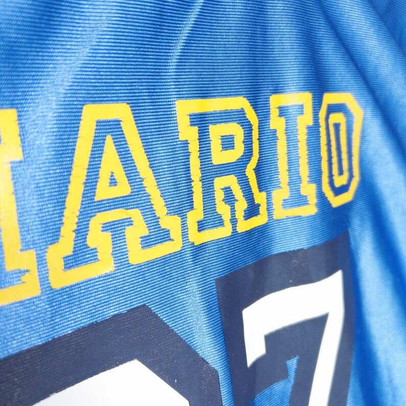 Puede incluir: Una camiseta deportiva de color azul brillante con el texto amarillo "ARIO" en fuente de bloque. Debajo del texto, un n&uacute;mero "7" grande de color blanco est&aacute; impreso en la tela. El material tiene una textura acanalada horizontal visible.