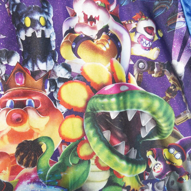 Puede incluir: Una tela con un fondo morado vibrante de tem&aacute;tica espacial que presenta un patr&oacute;n repetitivo de varios personajes de videojuegos, incluyendo una gran planta pira&ntilde;a verde con dientes blancos afilados, una criatura roja con corona y Bowser.