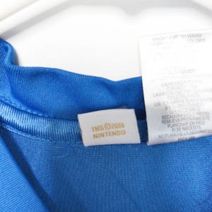 Puede incluir: Una camiseta de color azul brillante con una etiqueta interior blanca en el cuello con el texto dorado TM & &copy; 2008 NINTENDO y una etiqueta de cuidado m&aacute;s grande con instrucciones de lavado en ingl&eacute;s y espa&ntilde;ol para la prenda.