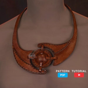 Könnte beinhalten: Eine braune Lederkette mit einem runden Design in der Mitte. Die Kette besteht aus zwei Lederstücken, die in der Mitte miteinander verbunden sind. Die Kette befindet sich an einem Mannequin-Hals.