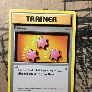Puede incluir: Una carta de comercio de Pokemon con una carta de entrenador llamada "Revive". La carta tiene un fondo plateado con un marco de madera. La carta muestra tres estrellas rosas con contornos amarillos. El texto de la carta dice "Pon un Pokémon Básico de tu pila de descarte en tu Banca."