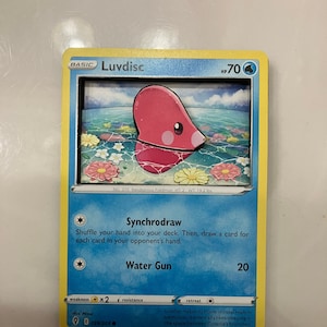 May include: A Luvdisc Pokemon card from the Sword & Shield - Vivid Voltage set. The card is number 039/203 and features a pink Luvdisc with a blue background. The card's text reads "Synchrodraw" and "Water Gun".