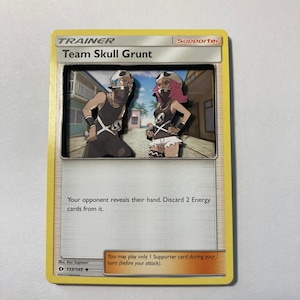 Puede incluir: Una carta de comercio de Pokémon con Team Skull Grunt de la expansión Sol y Luna. La carta es amarilla con texto negro y presenta una ilustración de dos miembros de Team Skull Grunt. El texto de la carta dice "Tu oponente revela su mano. Descarta 2 cartas de Energía de ella. Puedes jugar solo 1 carta de Partidario durante tu turno (antes de tu ataque)."