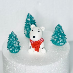 May include: A white polar bear cake topper wearing a red scarf, surrounded by three teal green frosted trees on a white cake.