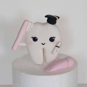 May include: A whimsical white tooth character with a graduation cap, holding a toothbrush and a diploma. A pink tube of toothpaste rests nearby. The tooth has a smiling face and is on a white foam base.