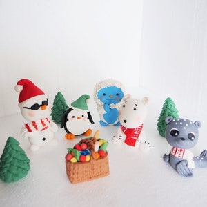 May include: A set of seven Christmas-themed fondant figurines, including a snowman wearing sunglasses, a penguin, a polar bear, a yeti, a seal, and two green Christmas trees. The figurines are arranged on a white surface.