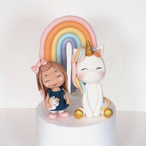 May include: A cake topper featuring a girl, a unicorn, and a rainbow. The girl wears a blue dress and holds a small unicorn. The unicorn has a golden horn and colorful mane. The rainbow has pastel colors.