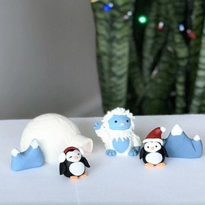 May include: A blue and white fondant snowman figurine, a white fondant igloo, and two penguins wearing red hats, all on a white surface.