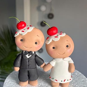 Puede incluir: Un par de adornos para pastel de bodas caprichosos. Una figura viste un esmoquin negro, la otra un vestido blanco. Ambos tienen glaseado con cerezas y se toman de la mano. Decoración de boda.