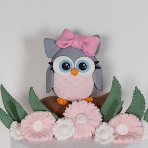 May include: A decorative owl figurine with a pink bow, perched on a brown branch. The owl has gray wings and a pink body, with large blue eyes. The base features pink and white flowers and green leaves.