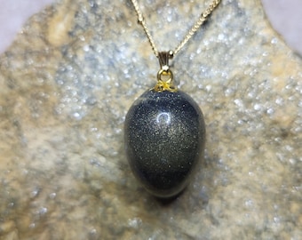 Op de afbeelding: Een goudkleurige ketting met een donkerblauwe, goudkleurige hanger. De eivormige hanger hangt aan een fijne ketting. De ketting wordt gepresenteerd op een getextureerd, lichtbruin stenen oppervlak.