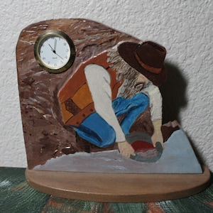 May include: A wooden clock featuring a painted scene of a prospector panning for gold. The clock has a round, gold-colored face with black numerals and hands. The prospector is wearing a brown hat, orange vest, and blue jeans. The clock is mounted on a wooden base.