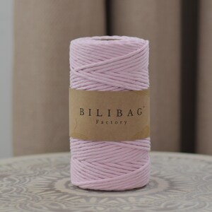 May include: A spool of light pink cotton macrame cord. The cord is tightly wound and wrapped with a brown paper label that reads "BILIBAG Factory". The cord is displayed on a decorative surface.