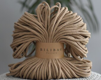 May include: A tightly wound coil of tan-colored macrame cord. The cord is wrapped around a cardboard label with the text "BILIBAG Factory" printed on it. The cord is resting on a gray, textured surface, with a blurred green plant in the background.