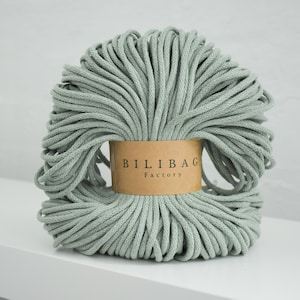 Op de afbeelding: Een bundel lichtgroen macramékoord. Het koord is strak opgerold en verpakt met een bruin papieren label met de tekst "BILIBAG Factory". Het koord ligt op een wit oppervlak.