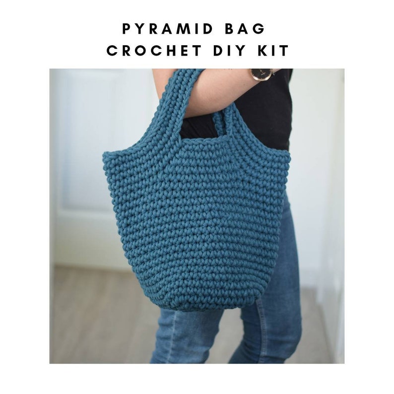 May include: A teal blue crocheted tote bag with a pyramid shape. The bag is being held by a person wearing jeans.