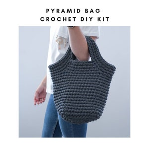 May include: A dark gray crocheted tote bag with a pyramid shape. The bag has a wide, flat base and a narrow top. The bag is being held by a person wearing a white shirt and blue jeans. The text "PYRAMID BAG CROCHET DIY KIT" is displayed above the image.
