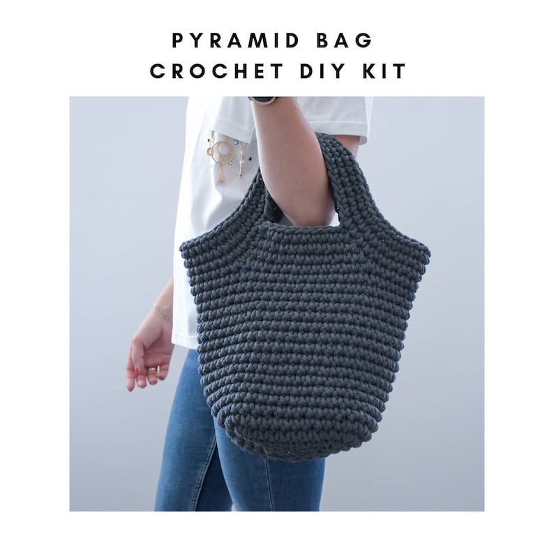 May include: A dark gray crocheted tote bag with a pyramid shape. The bag has a wide, flat base and a narrow top. The bag is being held by a person wearing a white shirt and blue jeans. The text "PYRAMID BAG CROCHET DIY KIT" is displayed above the image.