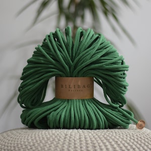 Pode incluir: Um feixe de corda de macramé verde, bem enrolada e macia ao toque, com uma etiqueta castanha com a inscrição "BILIBAG Factory". A corda é apresentada sobre um fundo desfocado. Uma pequena etiqueta está presa na extremidade da corda.