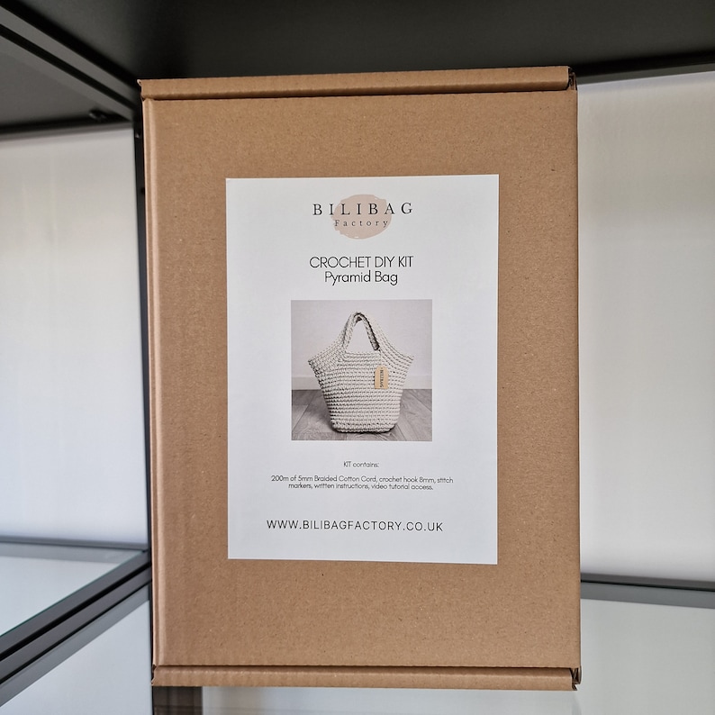May include: A brown cardboard box with a white label that says "BILIBAG Factory" and "CROCHET DIY KIT Pyramid Bag". The label also shows a picture of a white crocheted bag and the text "Kit contains 200m of 5mm Braided Cotton Cord, crochet hook, hook size, stitch marker, written instructions, video tutorial and access. WWW.BILIBAGFACTORY.CO.UK"