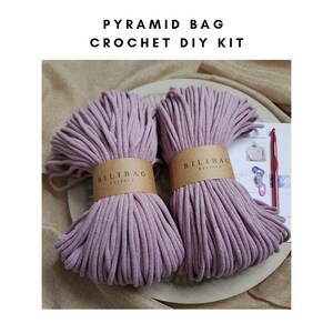 May include: A crochet DIY kit for making a pyramid bag. The kit includes two balls of purple cotton cord, a crochet hook, and a set of instructions. The cord is labeled "Bilibag Factory".