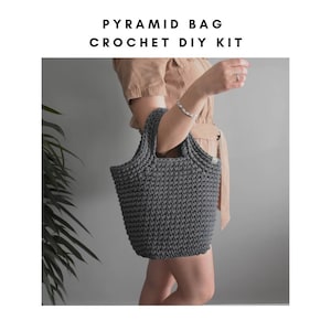 May include: A gray crocheted tote bag with a pyramid shape. The bag has two handles and is being held by a person.
