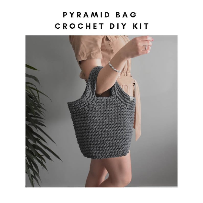 May include: A gray crocheted tote bag with a pyramid shape. The bag has two handles and is being held by a person.