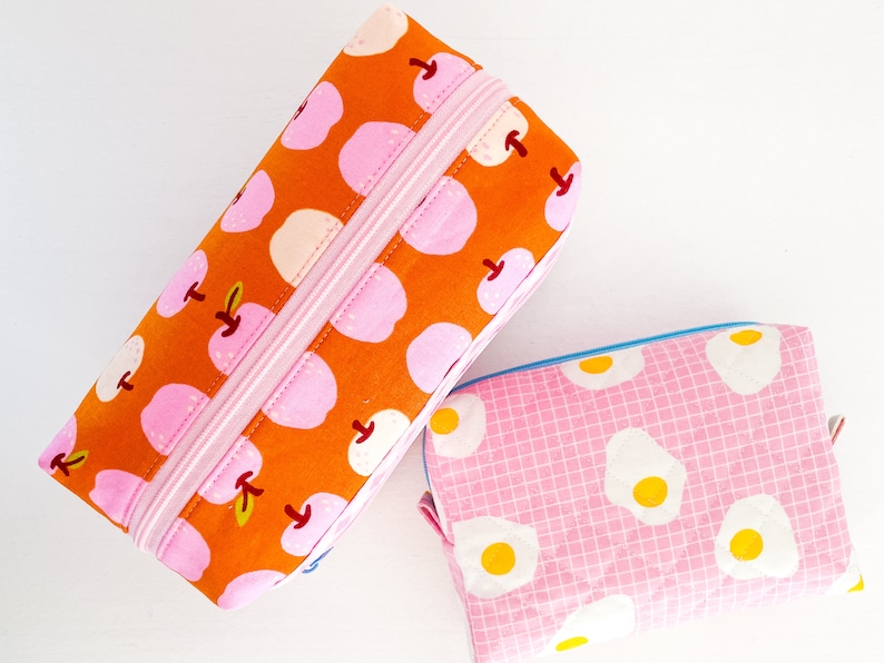 May include: Two fabric pencil cases. One is rectangular with an orange background and a pink apple pattern, and a pink zipper. The other is a smaller, rectangular pouch with a pink background and a fried egg pattern.