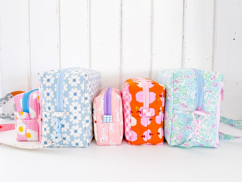 May include: A collection of colorful, patterned fabric zipper pouches. Designs include white daisies on blue, pink with white eggs, pink with white dogs, orange with pink apples, and floral patterns. Each pouch has a contrasting zipper.
