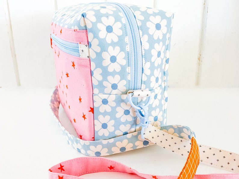 May include: A square crossbody bag with a light blue daisy print and a pink star pattern. The bag has a light blue zipper, a detachable strap, and a pink strap with red stars. The bag is approximately 8 inches wide.