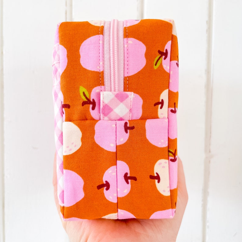 May include: A rectangular, zippered pouch with a repeating pattern of pink apples on an orange background. The zipper and handle are pink, and the side panel features a pink and white gingham pattern. The pouch is held in a hand.