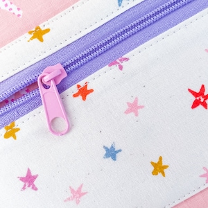 May include: Close-up of a pink and white fabric pencil case with a lavender zipper. The fabric is patterned with colorful stars. The zipper pull is pink.