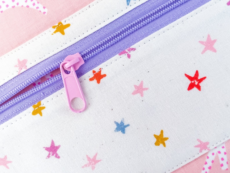May include: Close-up of a pink and white fabric pencil case with a lavender zipper. The fabric is patterned with colorful stars. The zipper pull is pink.