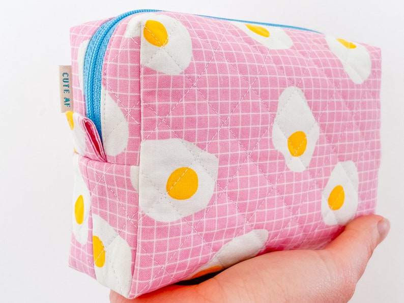 May include: A pink quilted cosmetic bag with a grid pattern and fried egg designs. The bag has a blue zipper and a small tag that reads "CUTE AF". The bag is rectangular and appears to be handmade.