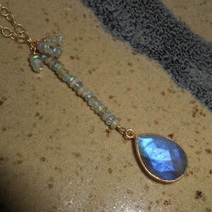 May include: A gold chain necklace with a teardrop-shaped blue labradorite gemstone pendant. The pendant is surrounded by a cluster of smaller, iridescent gemstones.
