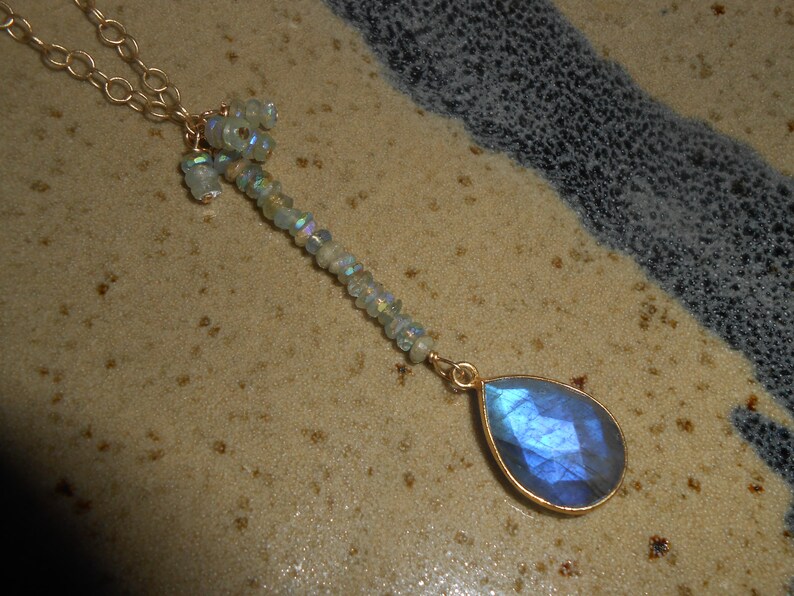May include: A gold chain necklace with a teardrop-shaped blue labradorite gemstone pendant. The pendant is surrounded by a cluster of smaller, iridescent gemstones.